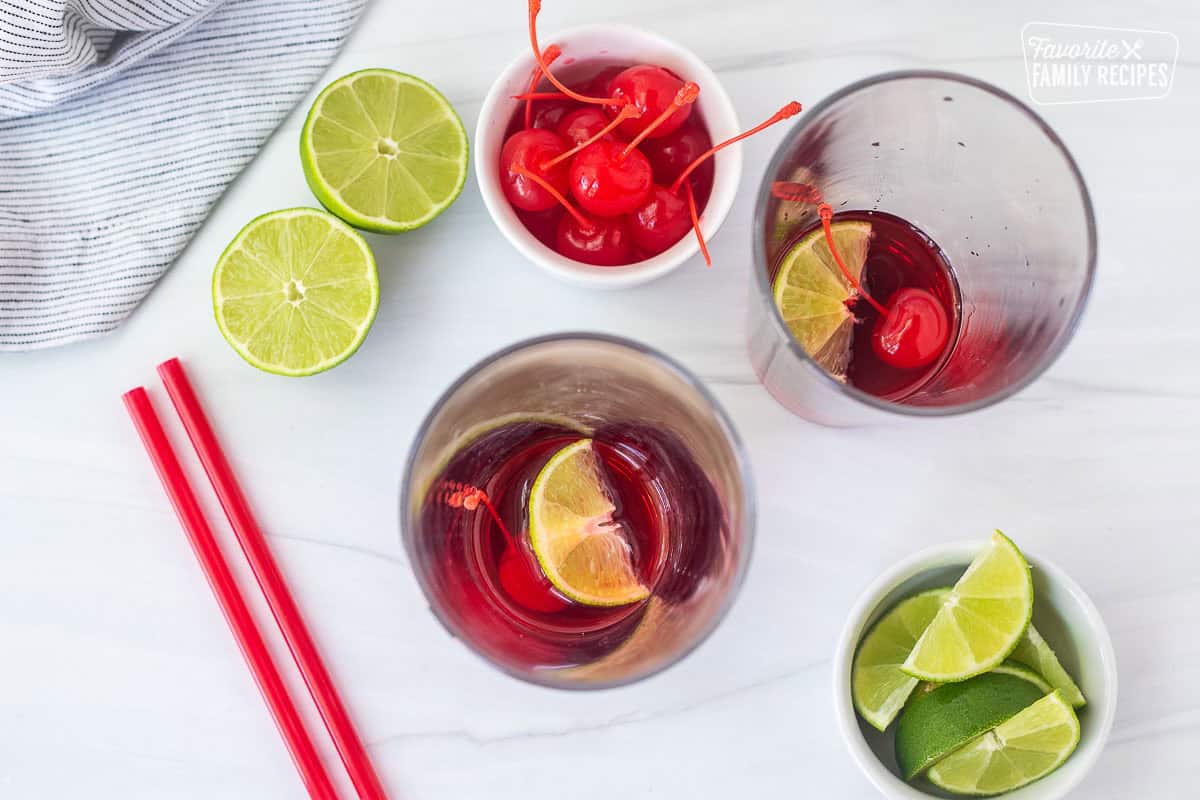 Two glasses with a lime wedge, cherry and cherry syrup on the bottom.