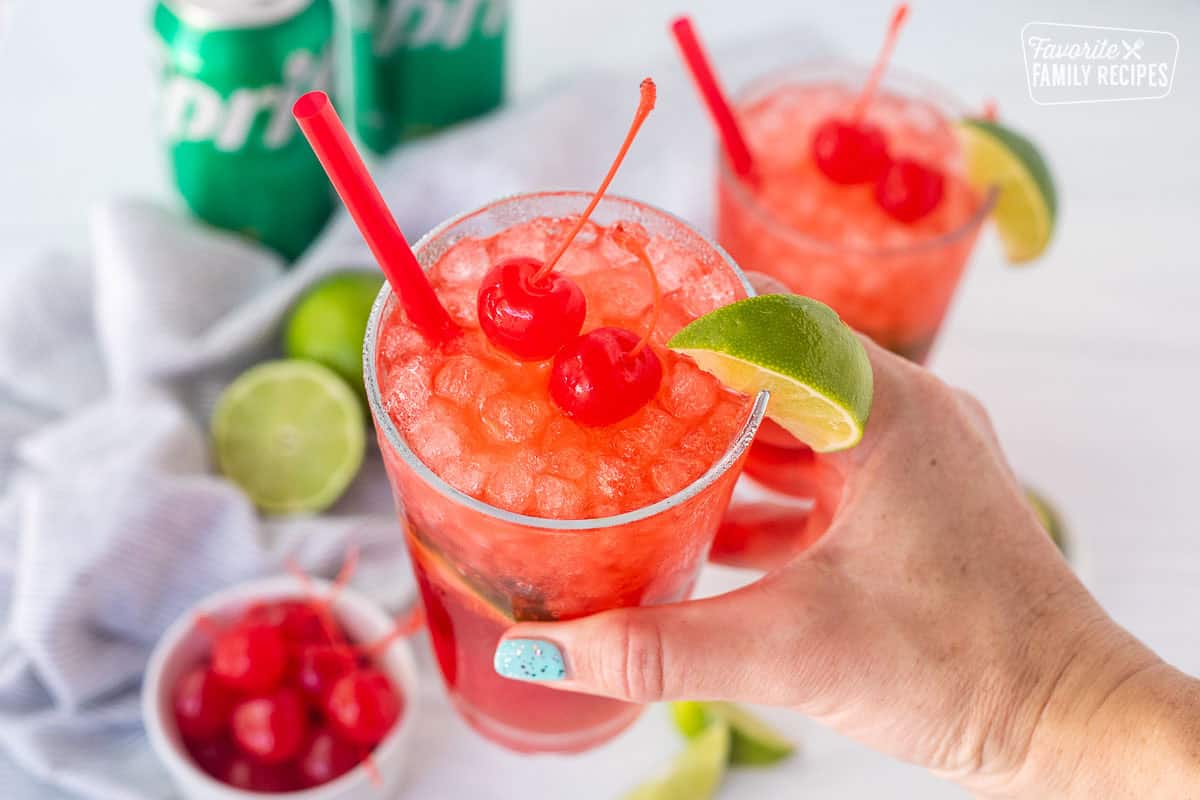 Hand holding a glass of cherry limeade and garnished with cherries and a lime wedge.
