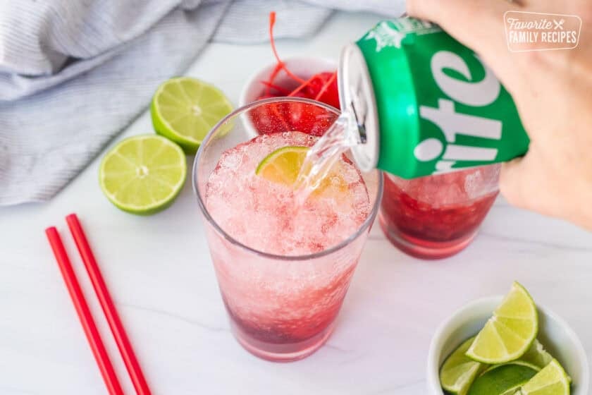 Pouring Sprite into glass with cherry syrup, ice, lime wedge and cherry.