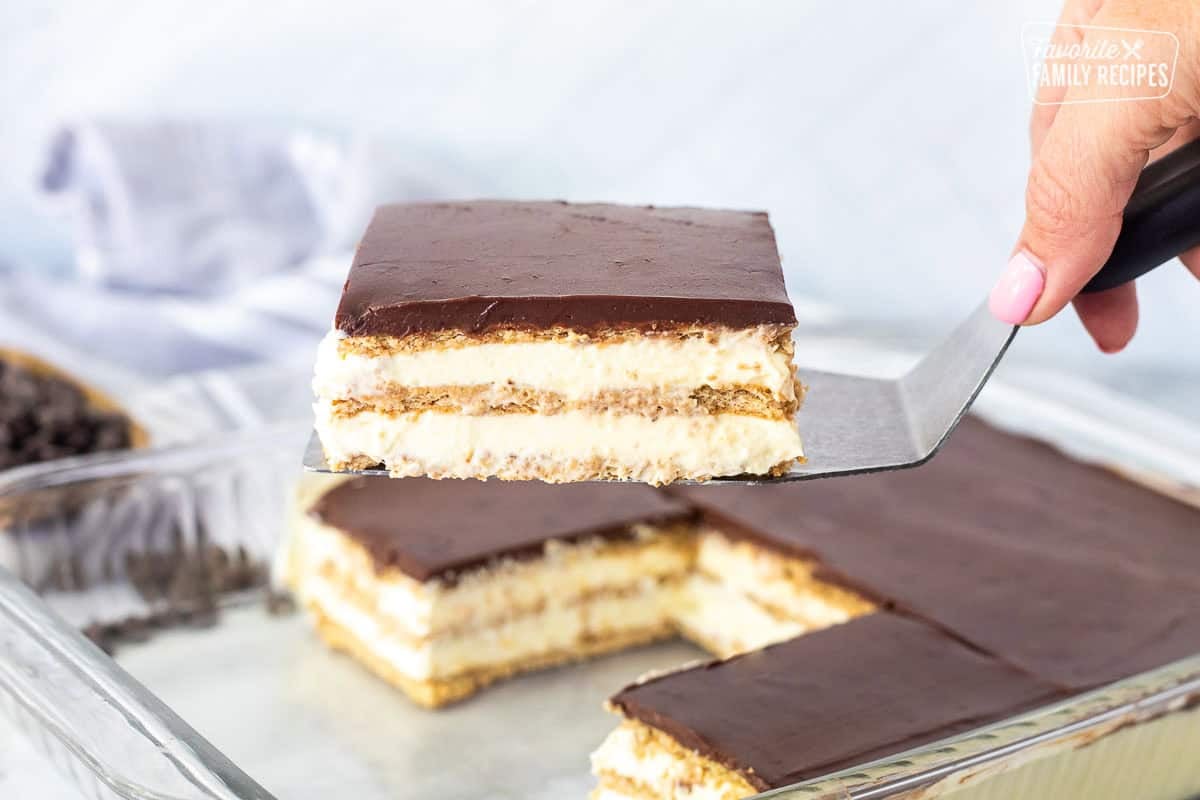 A hand holding a spatula with a slice of chocolate eclair cake with layers of graham crackers and pudding