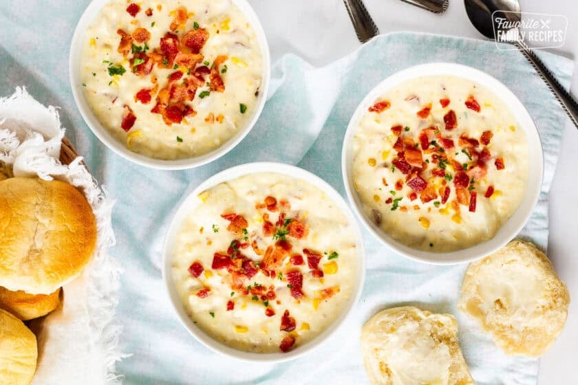 Three bowls of corn chowder with bacon topped with extra bacon and parsley. Dinner rolls on the side.