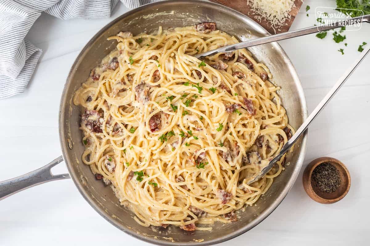Creamy carbonara in a skillet with tongs and garnished with parsley.