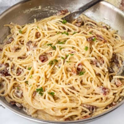 Creamy carbonara in a skillet with tongs and garnished with parsley.