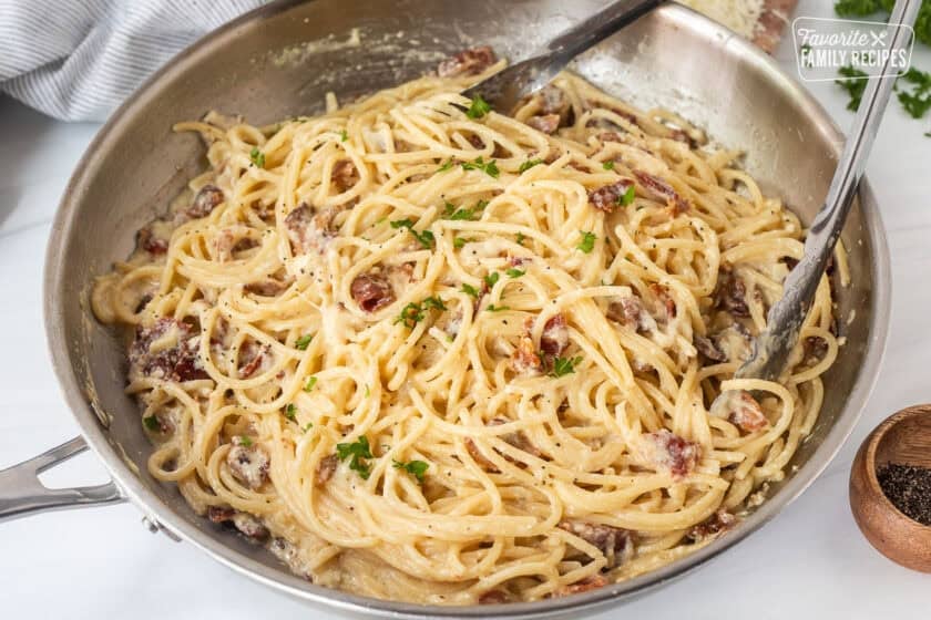 Creamy carbonara in a skillet with tongs and garnished with parsley.