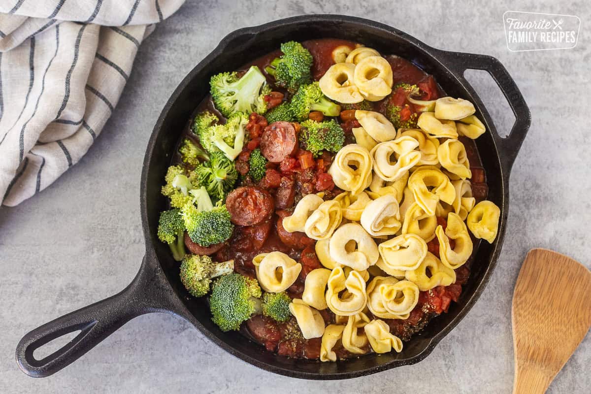 Adding frozen tortellini, tomato sauce, diced tomatoes, broccoli and spices to skillet of smoked sausage.