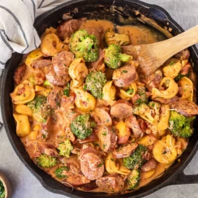 Skillet with creamy one pot tortellini with sausage and broccoli garnished with parsley and parmesan cheese. Wooden spoon on the side.