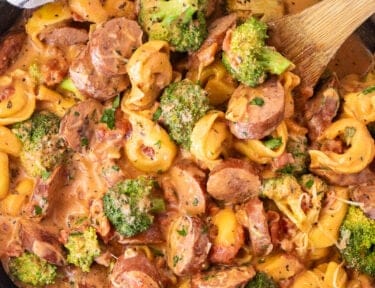 Skillet with creamy one pot tortellini with sausage and broccoli garnished with parsley and parmesan cheese. Wooden spoon on the side.