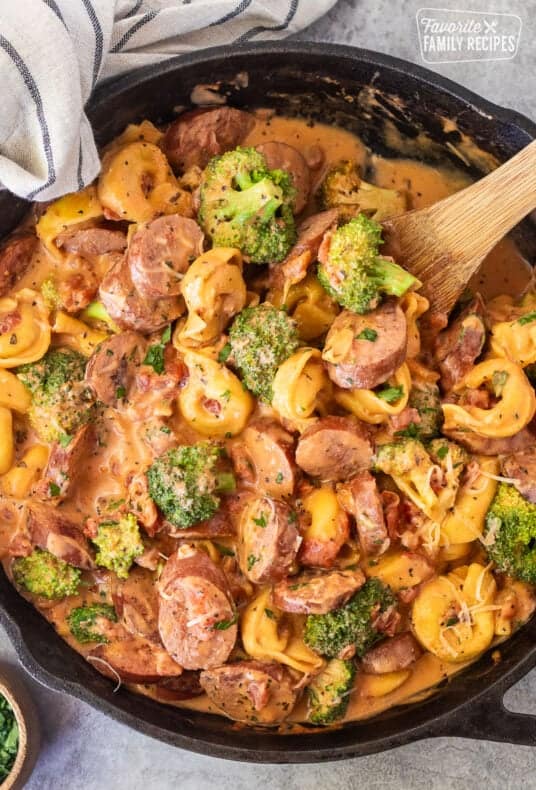 Skillet with creamy one pot tortellini with sausage and broccoli garnished with parsley and parmesan cheese. Wooden spoon on the side.