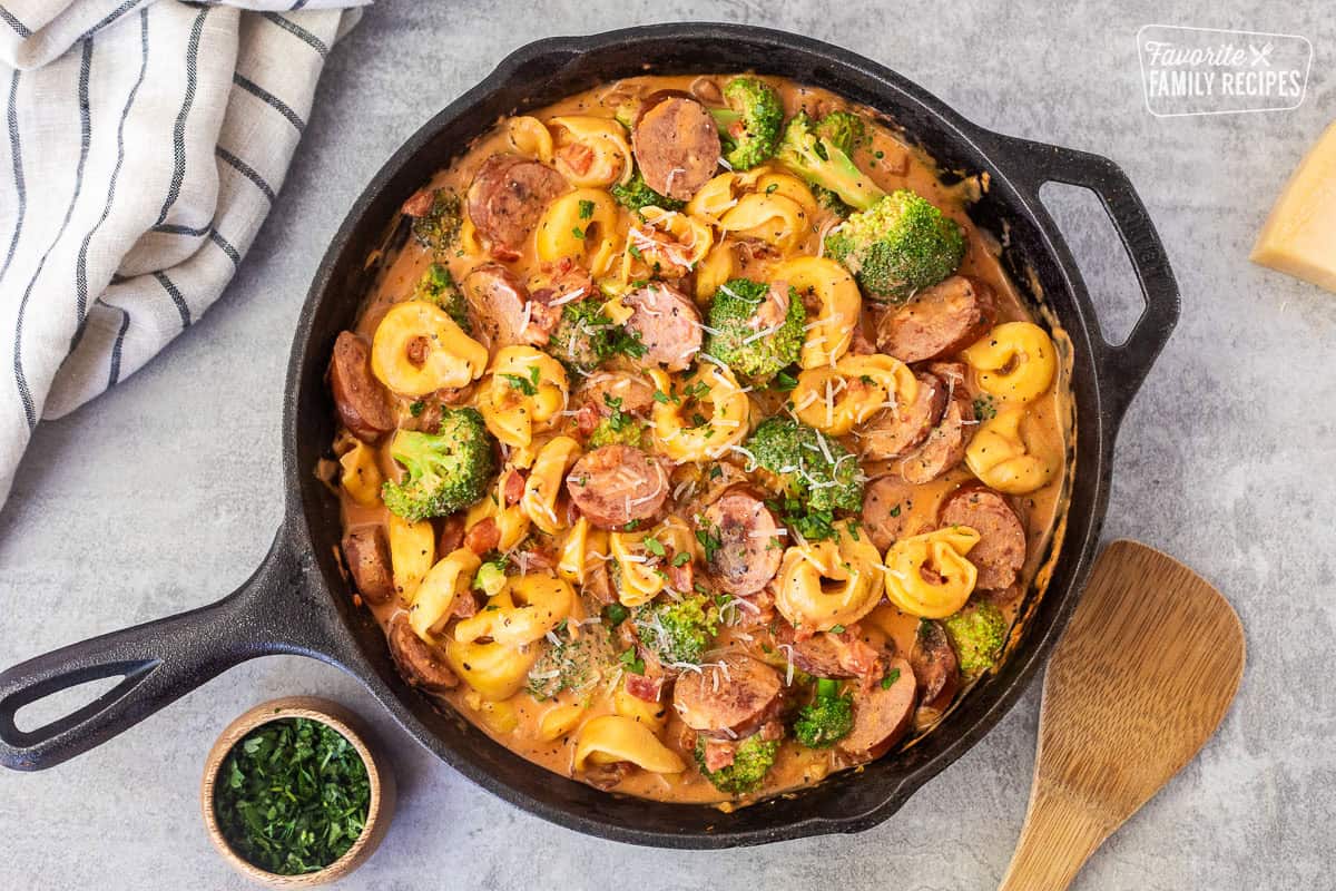 Skillet with creamy one pot tortellini with sausage and broccoli garnished with parsley and parmesan cheese. Wooden spoon on the side.