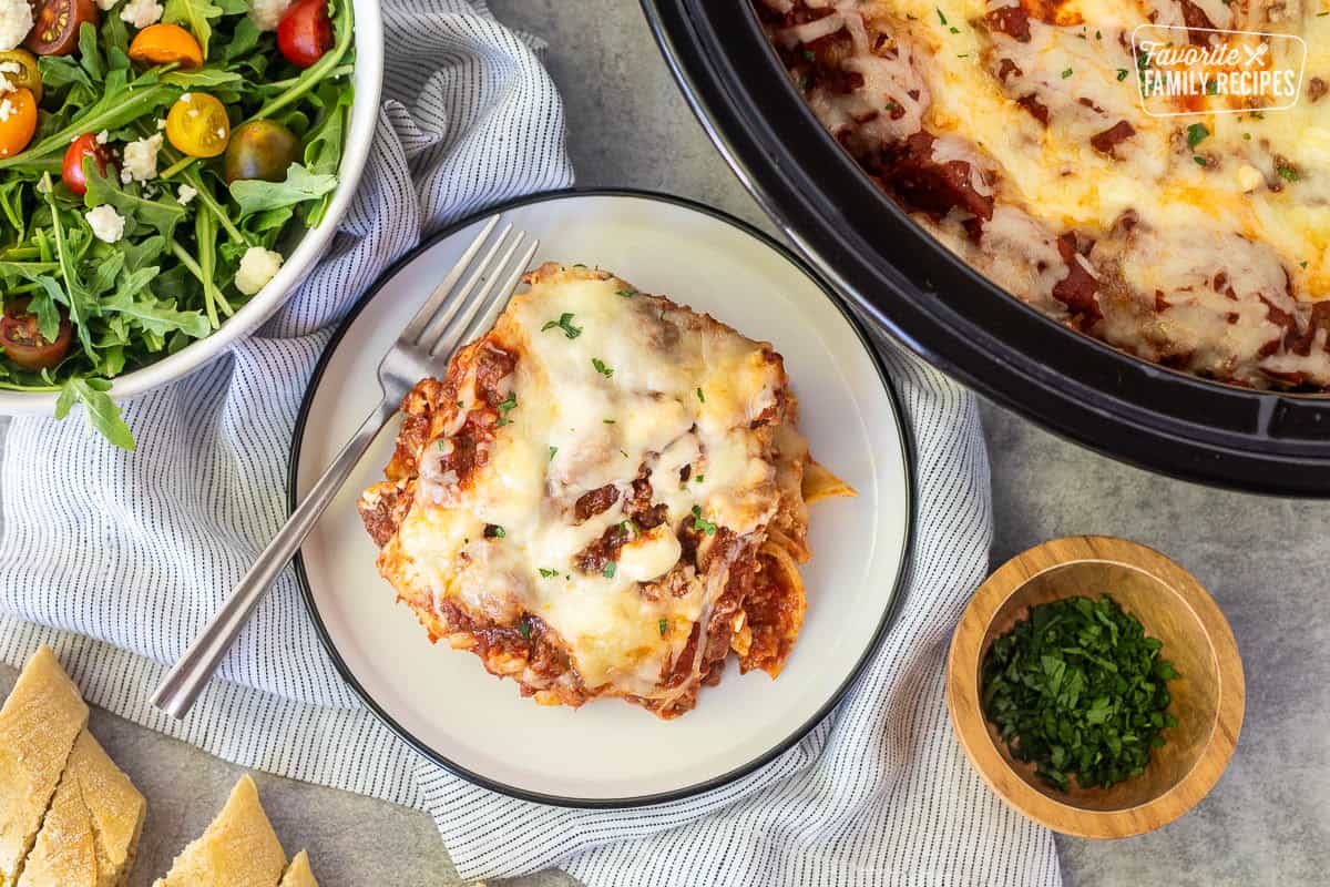 Slice of crockpot lasagna on a plate next to green salad and crockpot.