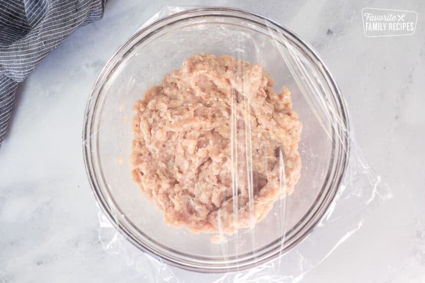 Glass mixing bowl with ground chicken mixture covered in plastic wrap.