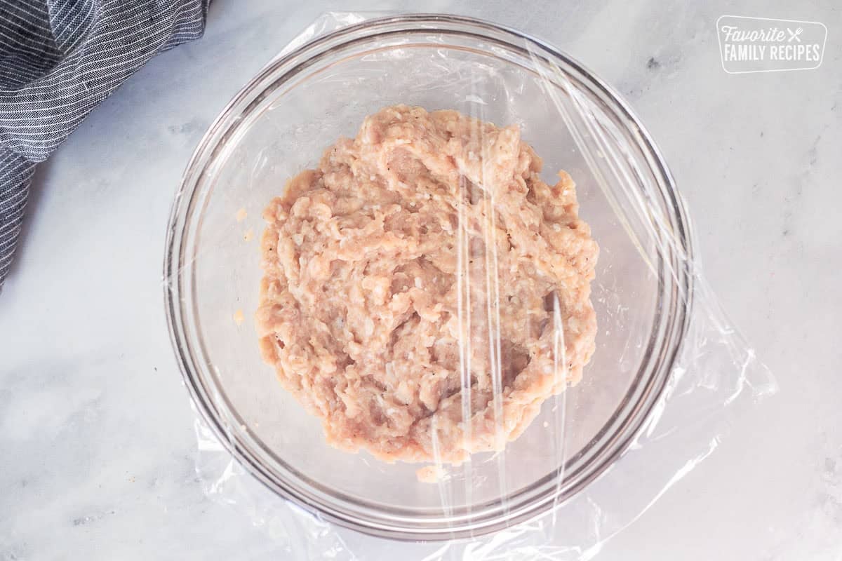 Glass mixing bowl with ground chicken mixture covered in plastic wrap.