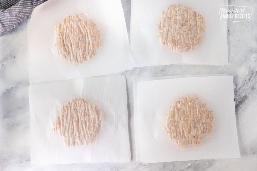 Four pieces of parchment paper with pressed down pieces of ground chicken patty mixture and a piece of parchment paper paper on top.