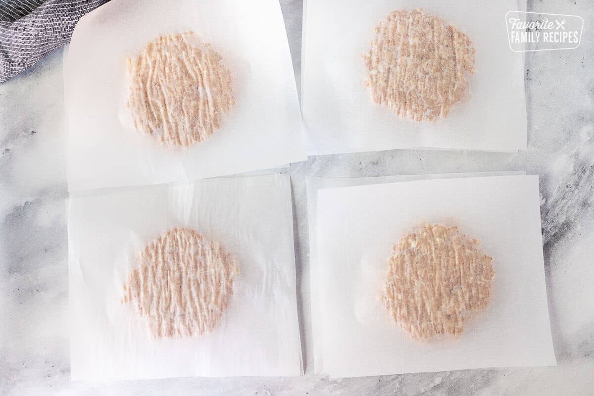 Four pieces of parchment paper with pressed down pieces of ground chicken patty mixture and a piece of parchment paper paper on top.