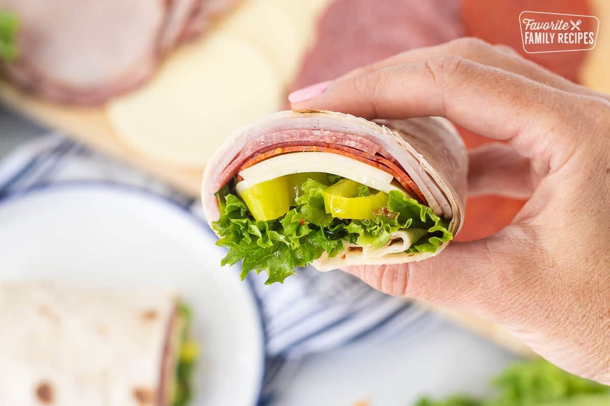 Hand holding an Italian wrap half with a flour tortilla, ham, salami, pepperoni, provolone cheese, lettuce, banana peppers and zesty Italian dressing.