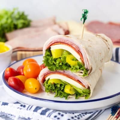 Italian wrap halves with a flour tortilla, ham, salami, pepperoni, provolone cheese, lettuce, banana peppers and zesty Italian dressing stacked together on a plate.