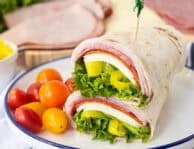 Italian wrap halves with a flour tortilla, ham, salami, pepperoni, provolone cheese, lettuce, banana peppers and zesty Italian dressing stacked together on a plate.