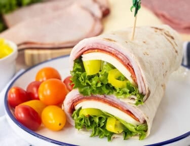 Italian wrap halves with a flour tortilla, ham, salami, pepperoni, provolone cheese, lettuce, banana peppers and zesty Italian dressing stacked together on a plate.