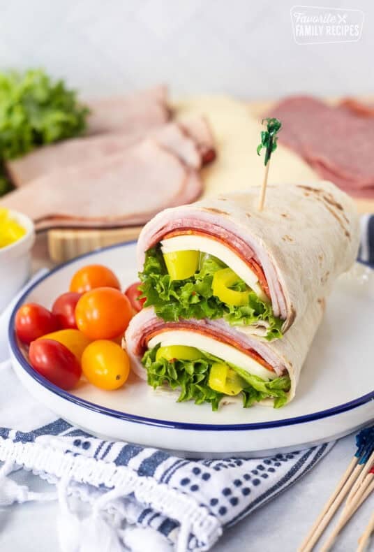 Italian wrap halves with a flour tortilla, ham, salami, pepperoni, provolone cheese, lettuce, banana peppers and zesty Italian dressing stacked together on a plate.
