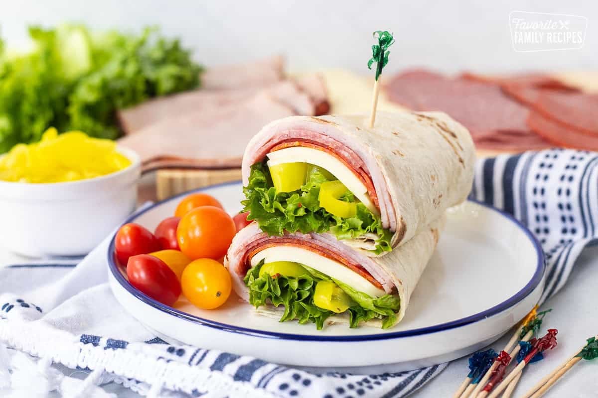 Italian wrap halves with a flour tortilla, ham, salami, pepperoni, provolone cheese, lettuce, banana peppers and zesty Italian dressing stacked together on a plate.