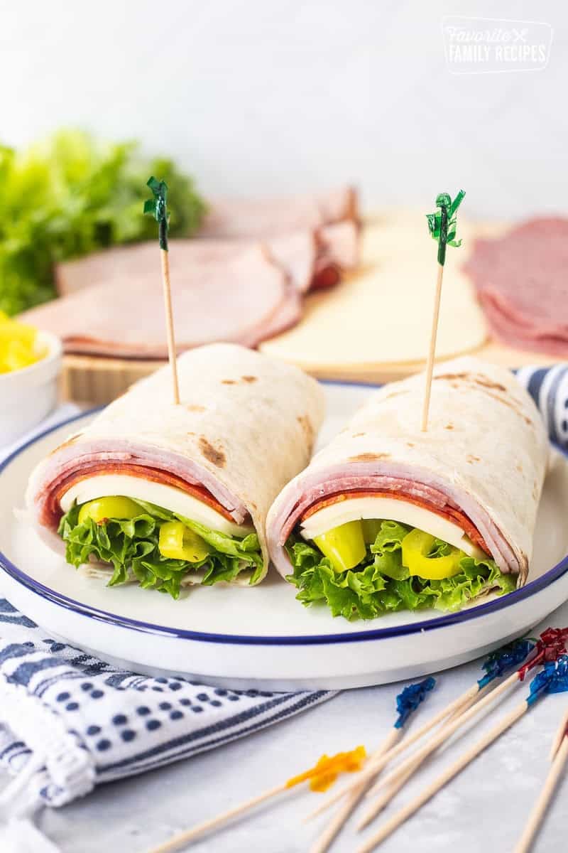 Italian wrap with a flour tortilla, ham, salami, pepperoni, provolone cheese, lettuce, banana peppers and zesty Italian dressing cut in half on a plate with toothpicks.