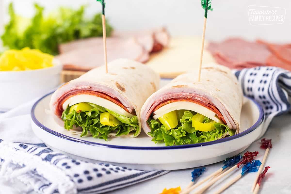 Italian wrap with a flour tortilla, ham, salami, pepperoni, provolone cheese, lettuce, banana peppers and zesty Italian dressing cut in half on a plate with toothpicks.
