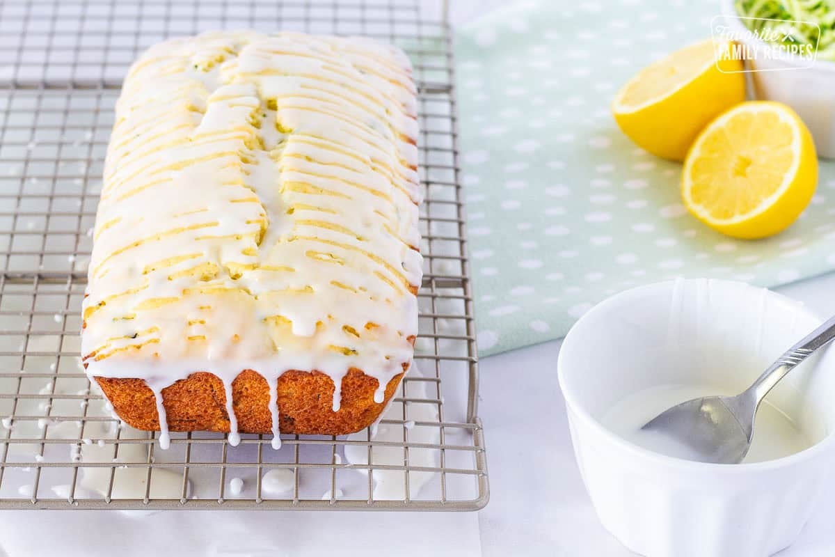 Drizzled lemon zucchini bread dripping from a cooling rack. Bowl and spoon of glaze on the side.