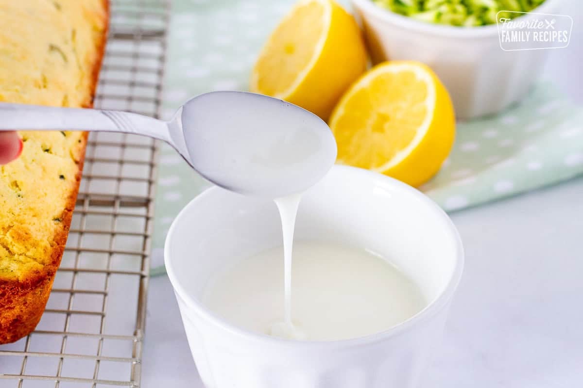 Lemon glaze drizzling from a spoon into a bowl for lemon zucchini bread.