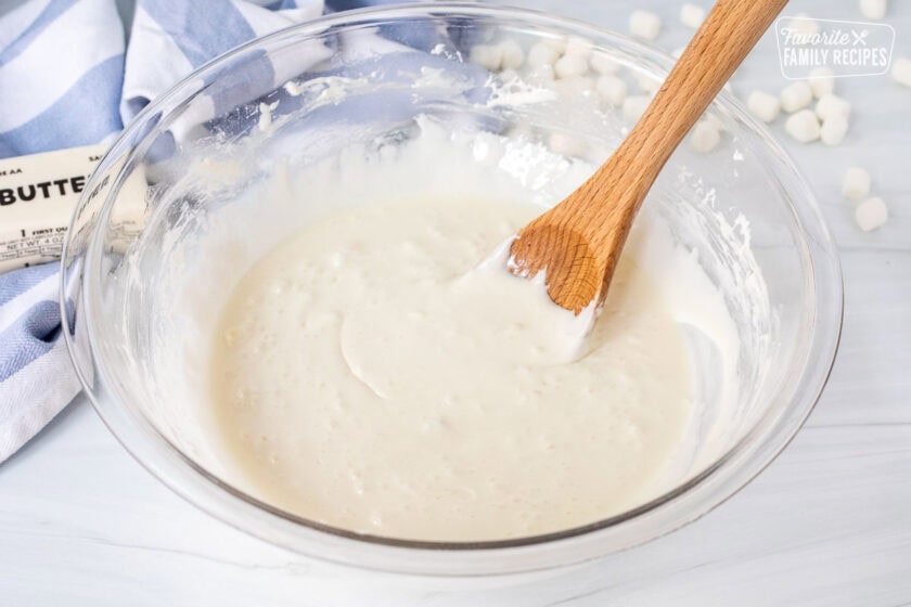 Glass mixing bowl of melted marshmallows and butter with a wooden spoon.