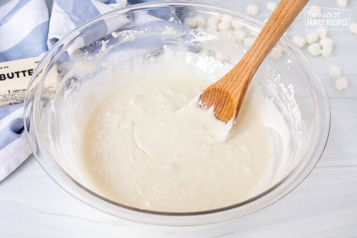 Glass mixing bowl of melted marshmallows and butter with a wooden spoon.