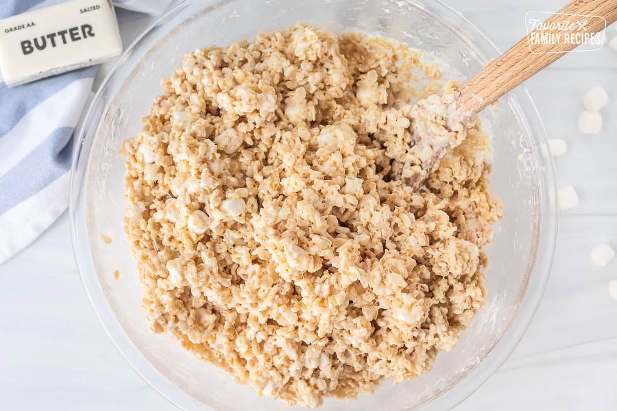 Glass mixing bowl with mixed rice krispie treats and wooden spoon.