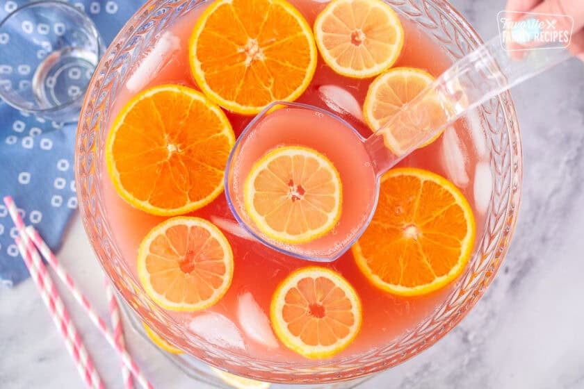 Ladle of party punch coming out of a large glass punch bowl with fresh lemon slices and orange slices.