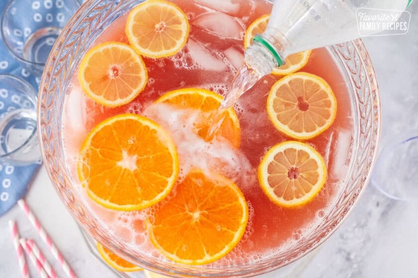 Pouring Sprite into large glass punch bowl of party punch with fresh slices of lemons and oranges floating on top.