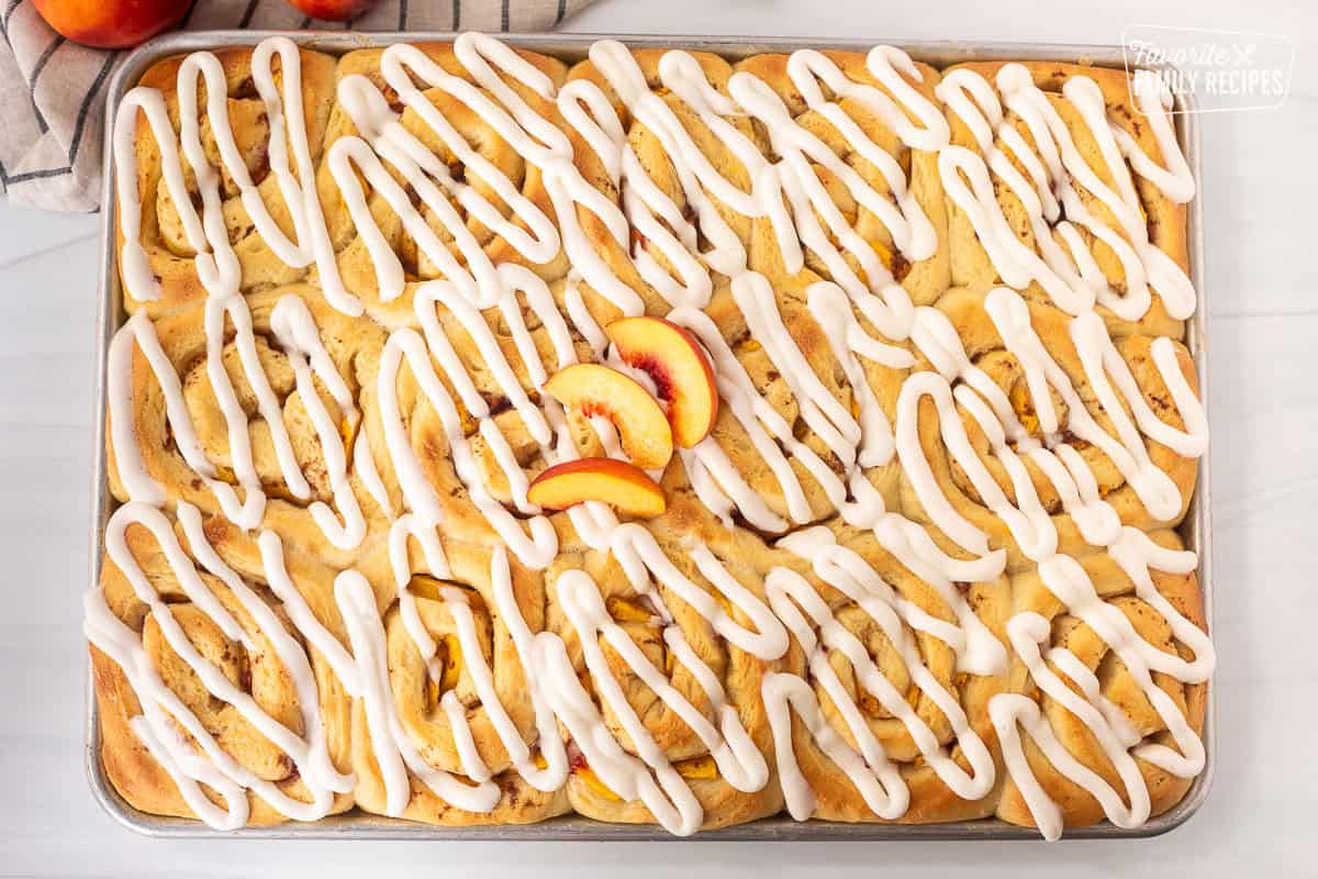 Frosting on top of peach cinnamon rolls in a sheet pan with sliced peaches.