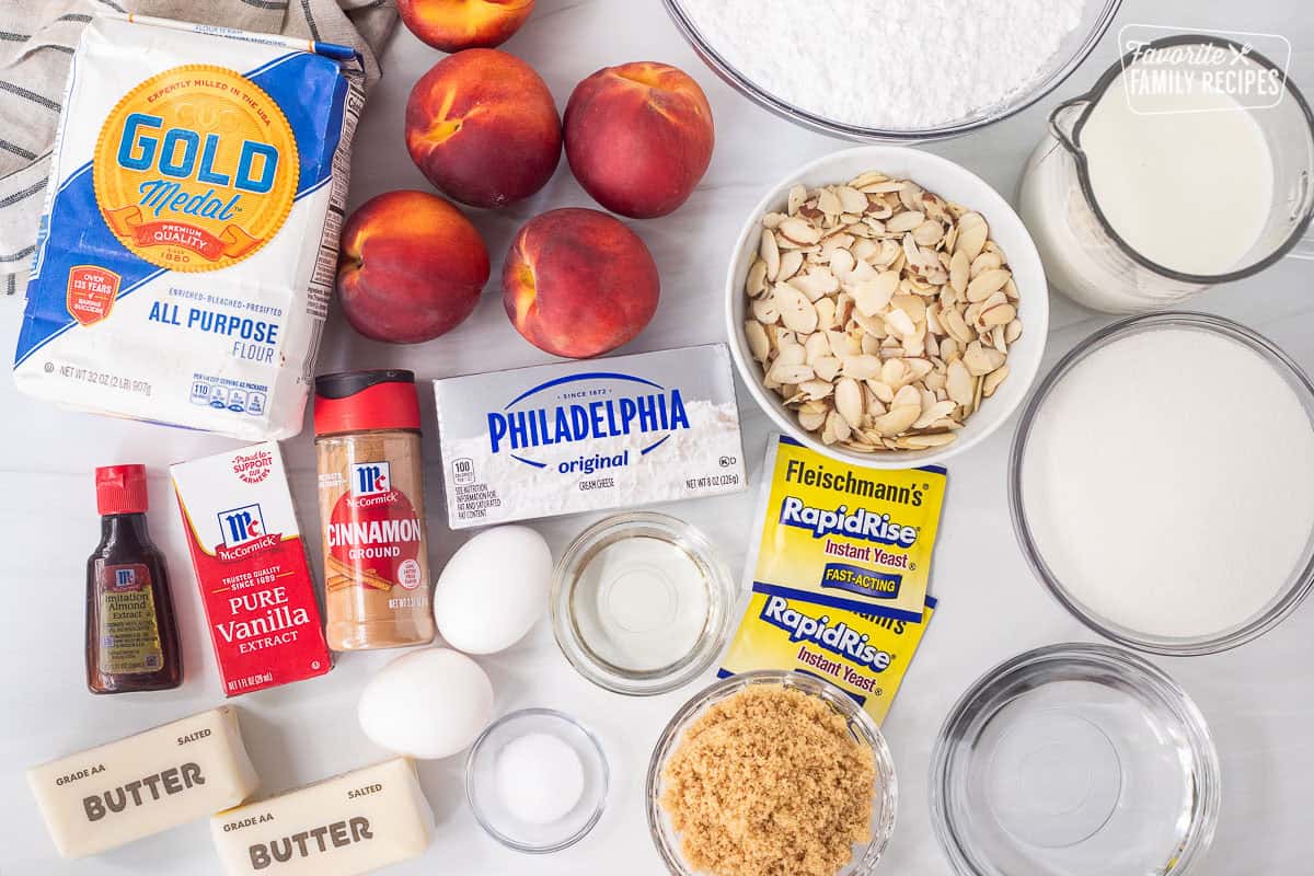 Ingredients to make peach cinnamon rolls including flour, peaches, sliced almonds, powdered sugar, milk, sugar, water, brown sugar, salt, oil, eggs, yeast, cream cheese, cinnamon, butter, almond extract and vanilla.
