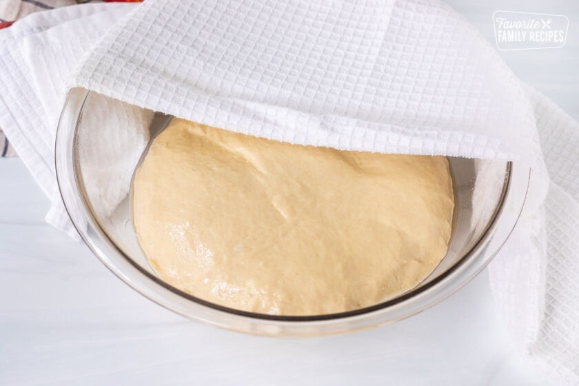 Towel over bowl of cinnamon roll dough ball rolled in oil.