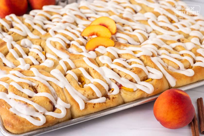 Frosting on top of peach cinnamon rolls in a sheet pan with sliced peaches.