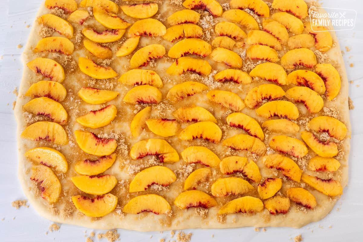 Rolled out cinnamon roll dough sprinkled with butter, sugars and cinnamon and sliced peaches.