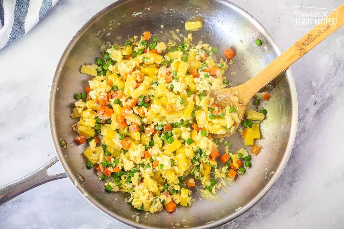 Cooked eggs mixed with pineapple and vegetables in a skillet with a wooden spoon.