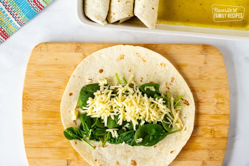 Cutting board with flour tortilla, spinach and cheese. Baking dish of enchiladas on the side.