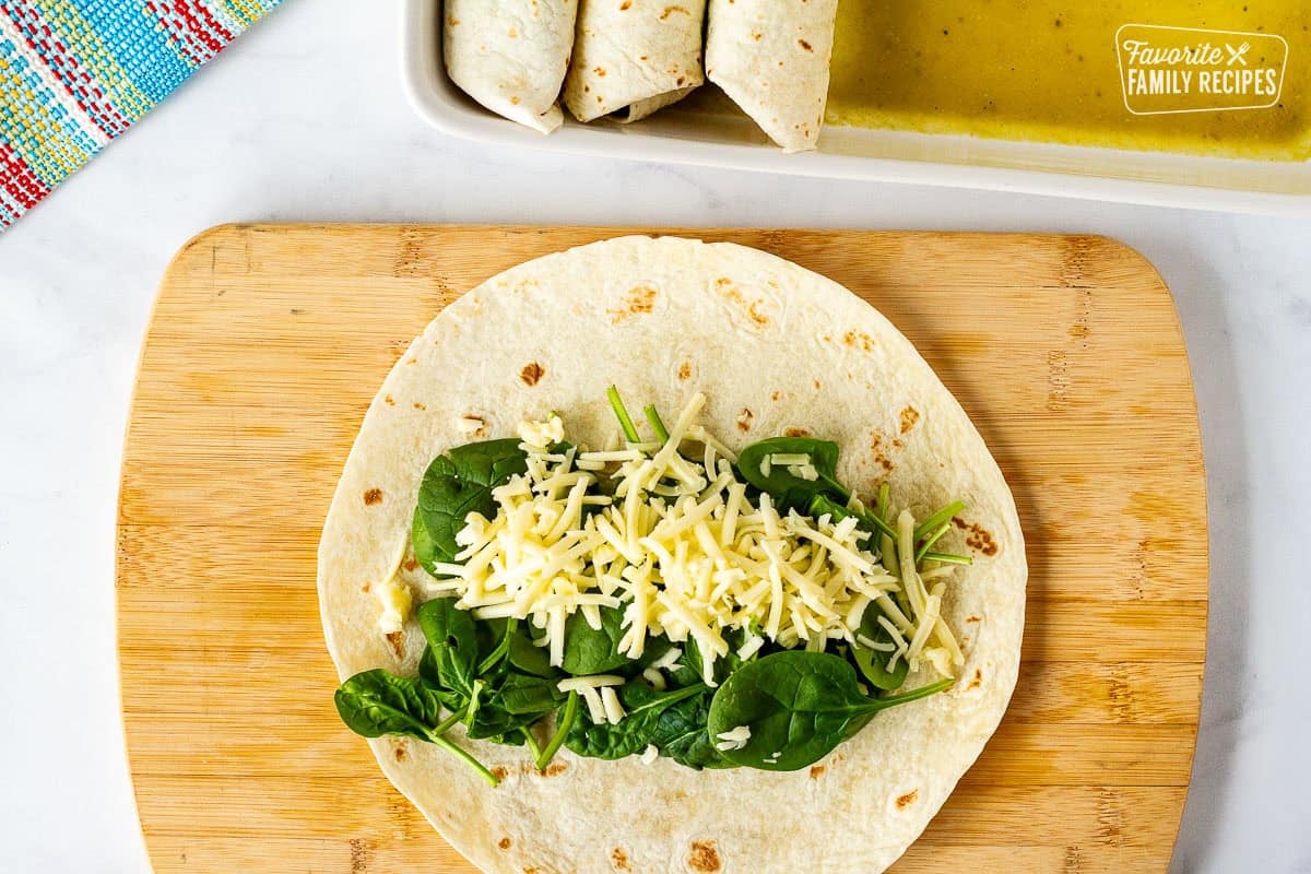Cutting board with flour tortilla, spinach and cheese. Baking dish of enchiladas on the side.