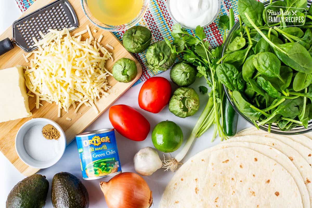 Ingredients to make Spinach Enchiladas including spinach, tomatillos, tortillas, green onion, cilantro, jalapeño, lime, garlic, onion, tomatoes, green chilies, avocado, cumin, salt, Monterey cheese, chicken broth, and sour cream.