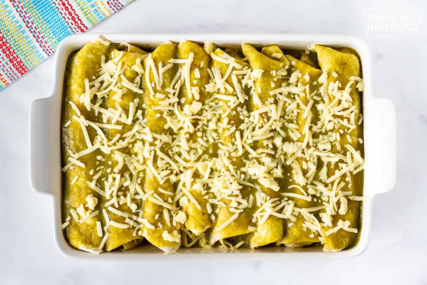Baking dish with unbaked rolled spinach enchiladas covered with green sauce and grated Monterey cheese.