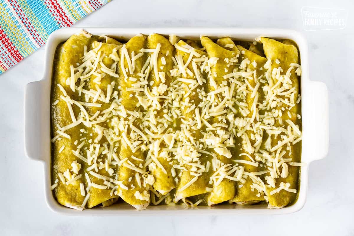 Baking dish with unbaked rolled spinach enchiladas covered with green sauce and grated Monterey cheese.