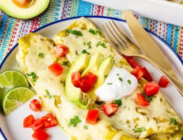 Plate with two green sauce spinach enchiladas garnished with sliced avocado, sour cream, tomatoes and cilantro.