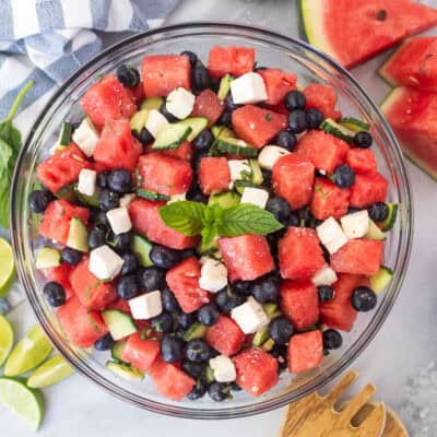 Watermelon feta salad in a glass bowl with watermelon, blueberries, feta cheese, cucumbers, milt, basil, salt and lime juice. Garnished with mint leaves.