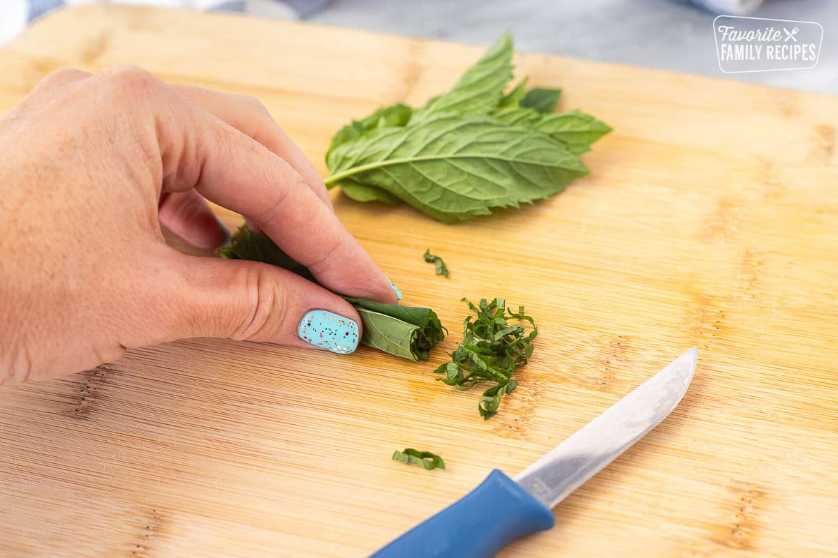 Cutting board with a hand curling the mint leaves and cutting in small strips with small knife.