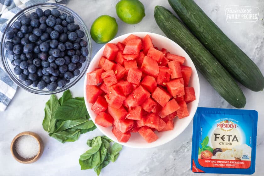 Ingredients to make watermelon feta salad including cucumbers, feta cheese, watermelon cubes, limes, blueberries, mint leaves, basil leaves, salt.