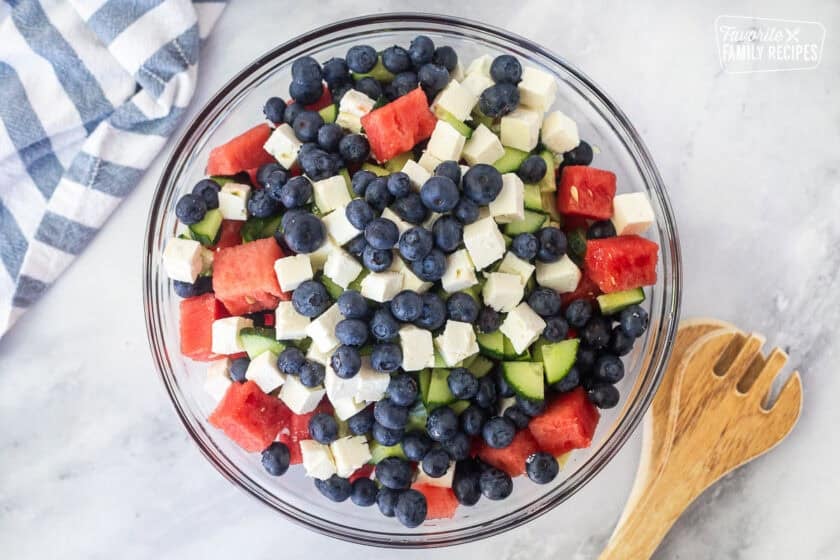 Glass bowl with watermelon, sliced cucumbers, blueberries and feta. Tongs on the side.