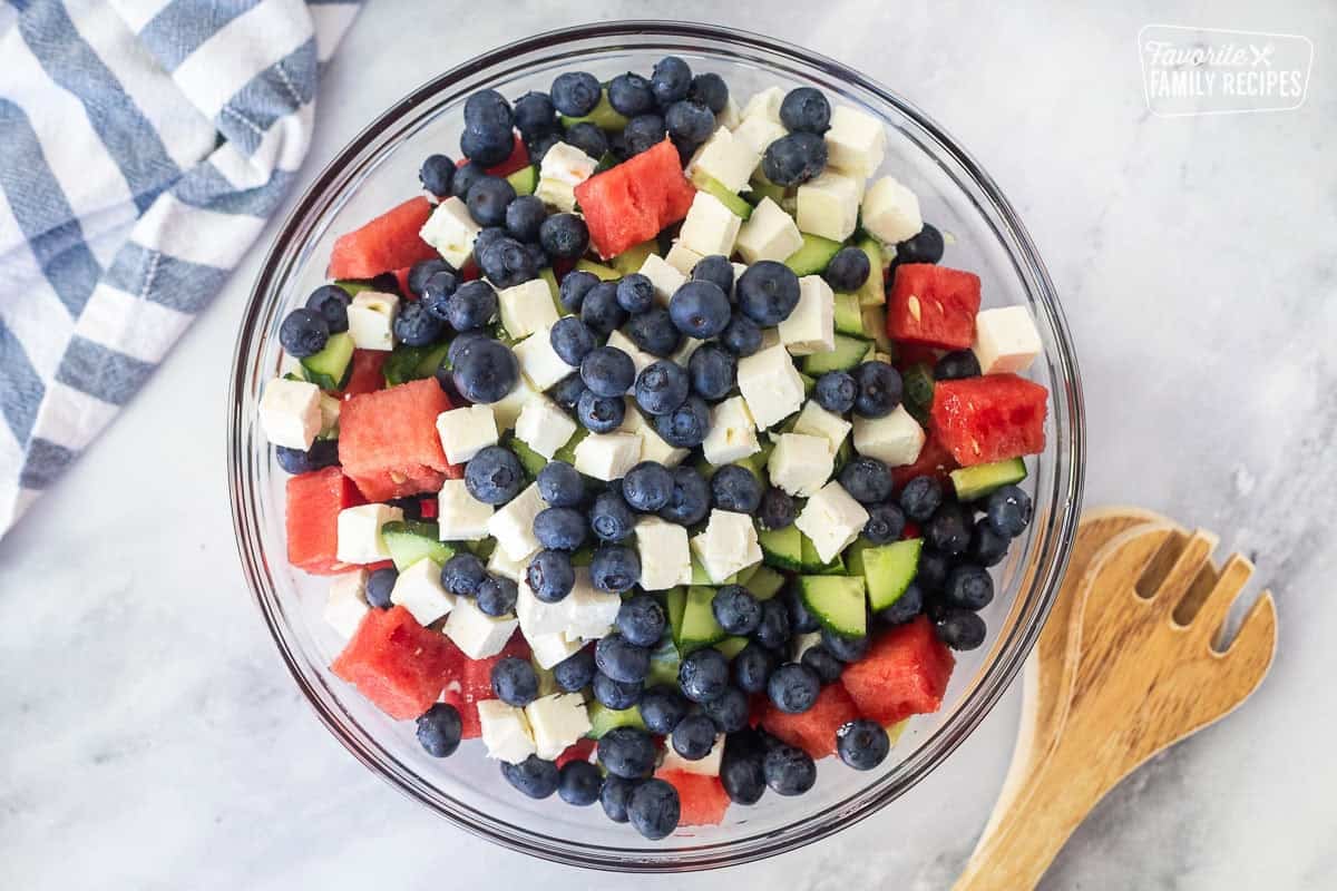 Glass bowl with watermelon, sliced cucumbers, blueberries and feta. Tongs on the side.